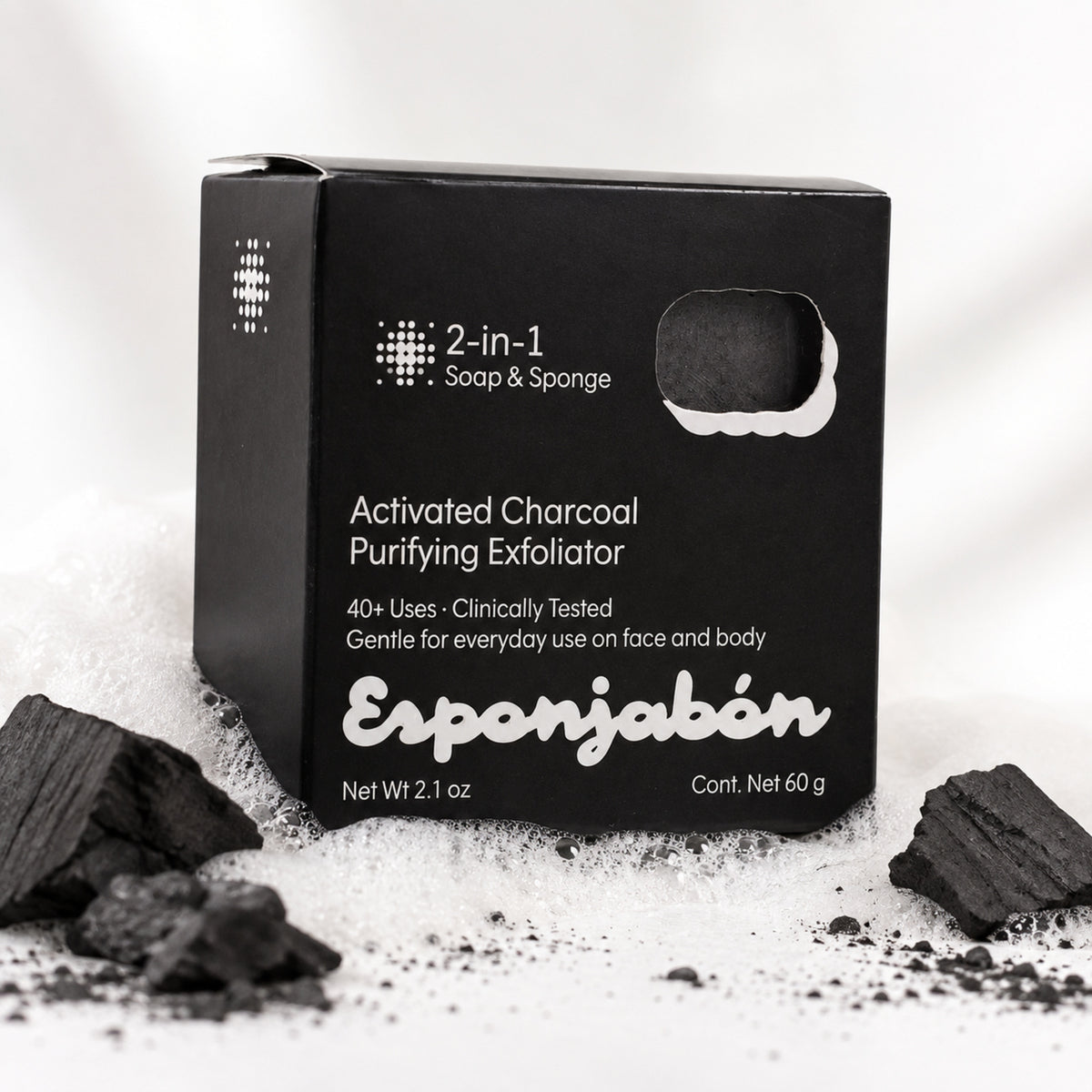 A black rectangular box of Esponjabén Activated Charcoal Purifying Exfoliator, surrounded by loose charcoal pieces and white powder on a white background.